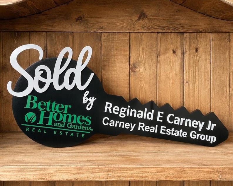 May include: A large black key-shaped sign featuring the white text Sold by, the green Better Homes and Gardens Real Estate logo, and the white text Reginald E Carney Jr Carney Real Estate Group printed across the blade of the key.