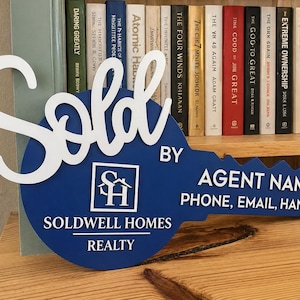 May include: A blue key-shaped sign with white lettering that reads Sold, Soldwell Homes Realty, Agent Name, Phone, Email, Handle, and features a square logo with the letters SH and a house icon in the center of the key head.