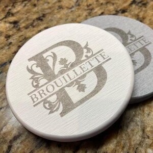 May include: Two circular white coasters with a brushed texture. Each coaster features a large, dark grey engraved letter B with ornate filigree details. The name BROUILLETTE is printed in a horizontal line across the center of the letter B.