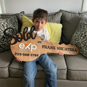 May include: A large, flat, wooden key-shaped sign featuring the black script text 'Sold', the white 'eXp REALTY' logo, and the black text 'BY FRANK NECATERA' with the phone number '203-568-3789' printed in black on the light brown wooden surface.