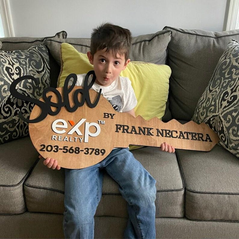 May include: A large, flat, wooden key-shaped sign featuring the black script text 'Sold', the white 'eXp REALTY' logo, and the black text 'BY FRANK NECATERA' with the phone number '203-568-3789' printed in black on the light brown wooden surface.
