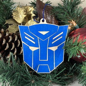 May include: A blue and natural wood Christmas ornament featuring the Autobots logo. The ornament is a geometric design with a blue background and natural wood accents. It has a blue string for hanging and is displayed among Christmas decorations.