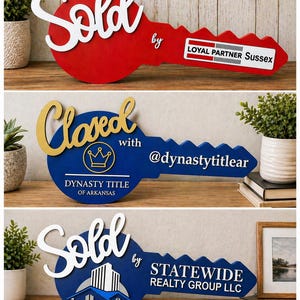 May include: Three decorative wooden key-shaped signs. The first is red with white text reading Sold by LOYAL PARTNER Sussex. The second is blue with gold text reading Closed with @dynastytitlear and DYNASTY TITLE OF ARKANSAS. The third is blue with white text reading Sold by STATEWIDE REALTY GROUP LLC.