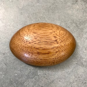 Puede incluir: Un cuenco ovalado de madera con una superficie pulida. El cuenco está hecho de madera marrón claro con vetas marrones oscuras. Está vacío y descansa sobre una superficie gris.