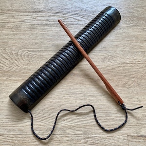 Puede incluir: Un instrumento de percusión guiro de color marrón oscuro con una superficie estriada y un raspador de madera. El guiro es cilíndrico con un cordón negro y naranja adjunto. El raspador es una fina varilla de madera marrón.