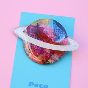 May include: A planet-shaped brooch with a white ring, set on a light blue card against a pink backdrop. The planet is a mix of red, blue, and yellow. The word "Peco" is visible on the card.