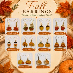 May include: A collection of ten pairs of gold-toned dangle earrings displayed on white cards, featuring autumn-themed enamel charms including maple leaves, owls, acorns, mugs, coffee cups, trees, pumpkins, sweaters, gourds, and snails.