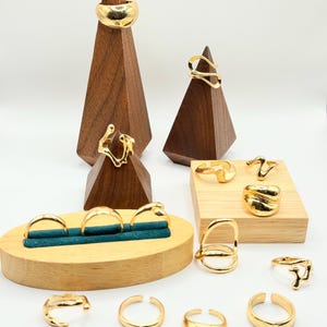 Può includere: Una collezione di anelli color oro in vari design, esposti su supporti in legno. Gli anelli includono fasce spesse, design aperti e forme curve. I supporti in legno hanno la forma di piramidi e blocchi, con un espositore in velluto verde acqua.