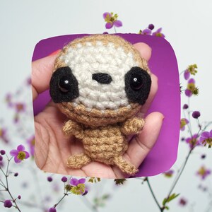 May include: A small crocheted sloth toy with brown and white fur, black eyes, and a black nose. The sloth is being held in a hand against a purple background.