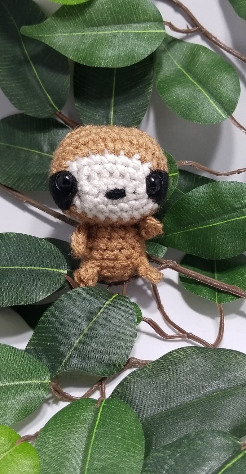 May include: A small crocheted sloth toy with brown fur, white face, and black eyes. The sloth is sitting on a branch with green leaves.