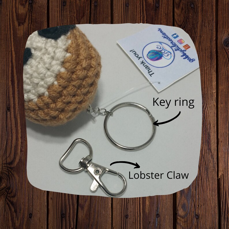 May include: Crocheted brown and white character with black eyes attached to a silver key ring with a lobster claw clasp. The key ring is a circle and the lobster claw is a hook.