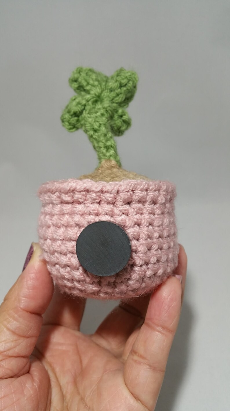 May include: A small crocheted potted plant with a green palm tree and a pink pot. The pot has a black circle on the back.