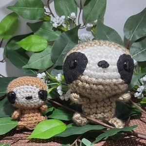 May include: Two crocheted sloth plushies, one brown and one beige, with black eyes and a white nose. The plushies are sitting on a brown woven mat with green leaves behind them.