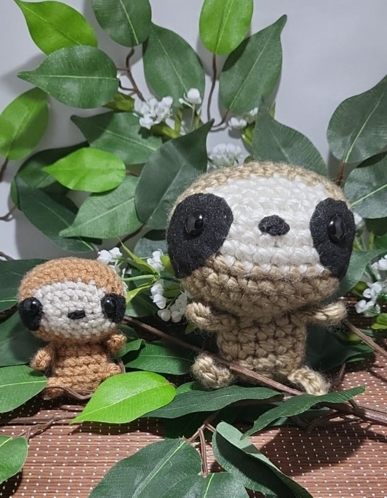 May include: Two crocheted sloth plushies, one brown and one beige, with black eyes and a white nose. The plushies are sitting on a brown woven mat with green leaves behind them.