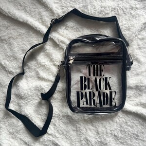 Puede incluir: Un bolso cruzado transparente con una correa y cremallera negras. El bolso presenta el texto "THE BLACK PARADE" en letras negras en negrita. El bolso es rectangular y tiene un ribete negro.