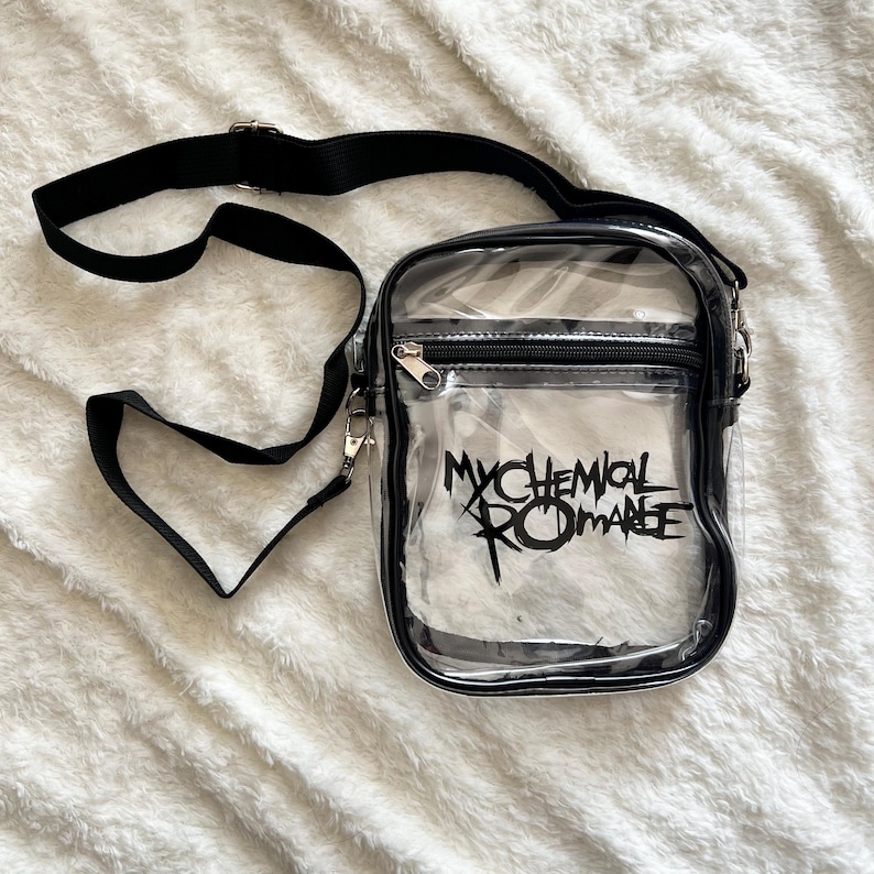 Puede incluir: Bolso bandolera transparente con ribete y correa negros. El bolso tiene un bolsillo frontal con cremallera y el texto "MY CHEMICAL ROMANCE" en negro. El bolso est&aacute; sobre una superficie blanca texturizada.
