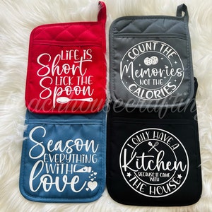 May include: Set of four red, gray, blue, and black oven mitts with white text. The mitts feature various kitchen-themed sayings, including "Life is Short Lick the Spoon", "Count the Memories Not the Calories", "Season Everything with Love", and "I Only Have a Kitchen Because It Came with the House".