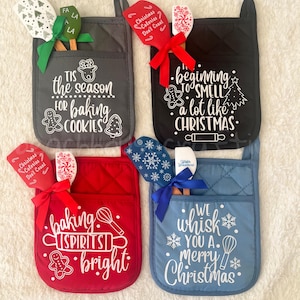 May include: Four quilted pot holders in gray, black, red, and blue, each with a pocket holding a wooden spoon and spatula. The pot holders have festive text like "Tis the season for baking cookies" and "We whisk you a Merry Christmas."