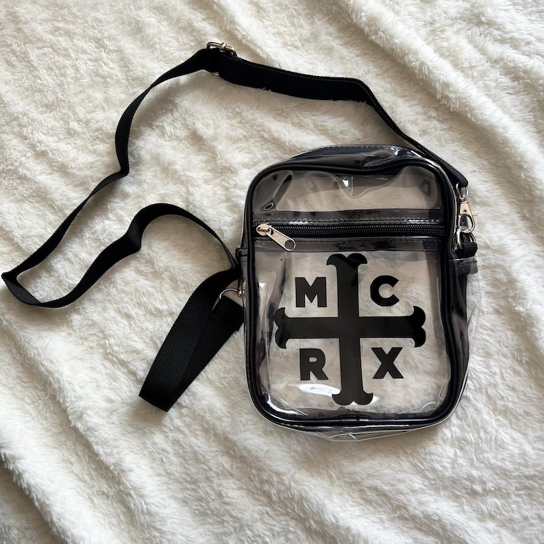 Puede incluir: Un bolso cruzado transparente con una correa y cierre de cremallera negros. El bolso presenta las letras "MCRX" en un dise&ntilde;o de cruz negra en la parte delantera. El bolso est&aacute; hecho de un material transparente.