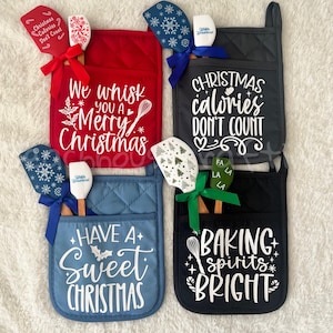 May include: Four Christmas-themed oven mitts in red, blue, black, and gray, each with a unique holiday message in white. Each mitt has a matching spatula with a festive design and a decorative ribbon.