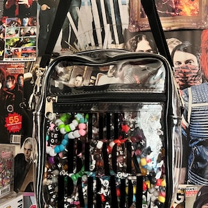 Puede incluir: Bolso bandolera transparente con correa y cremallera negras. La bolsa est&aacute; llena de cuentas de colores y las palabras "THE BLACK PARADE" en letras negras grandes. Carteles en el fondo.