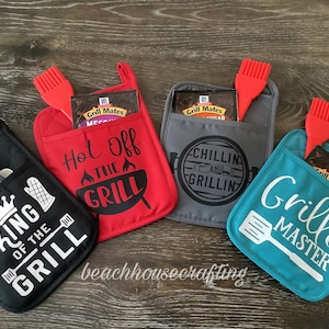 May include: Four different oven mitts with pockets for grilling tools and spice packets. The mitts are red, black, gray, and teal. The text on the mitts reads "Hot Off the Grill", "King of the Grill", "Chillin and Grillin", and "Grill Master".
