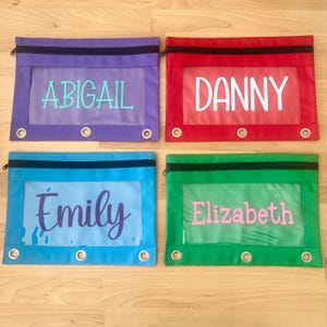 May include: Four colorful pencil cases with clear windows and personalized names: Abigail, Danny, Emily, and Elizabeth. Each case has a zipper closure and reinforced grommets for binder use. The cases are in purple, red, blue, and green.