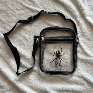 Puede incluir: Un bolso cruzado transparente con correa y ribete negros. El bolso presenta un gr&aacute;fico de ara&ntilde;a negra en la parte delantera. El bolso tiene una cremallera y un cierre de metal plateado.