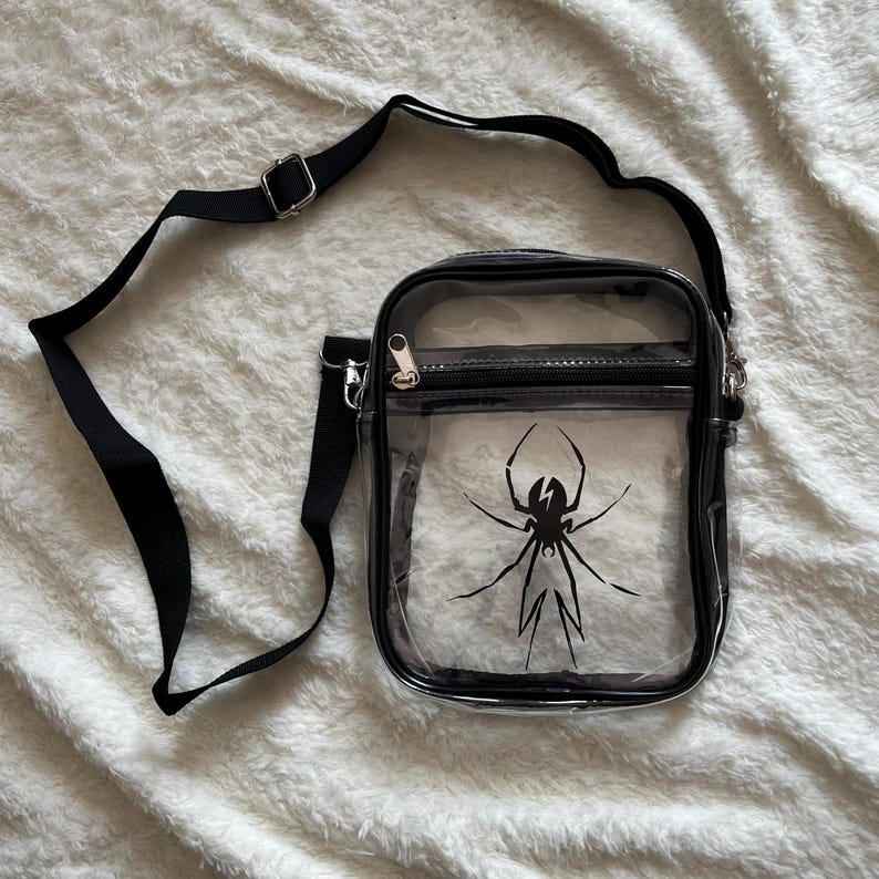 Puede incluir: Un bolso cruzado transparente con correa y ribete negros. El bolso presenta un gr&aacute;fico de ara&ntilde;a negra en la parte delantera. El bolso tiene una cremallera y un cierre de metal plateado.