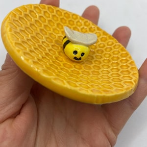 May include: A yellow ceramic soap dish with a honeycomb design. A small, smiling bee figurine with a white hat is centered on the dish. The dish is oval-shaped and held in a hand.