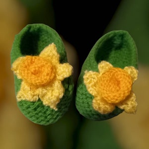 May include: A pair of green knitted baby booties with yellow daffodil flowers on the toes.