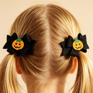 May include: A pair of black hair bows, each featuring a centred orange jack-o'-lantern pumpkin appliqué with a green stem. The bows are constructed from a solid black fabric and are attached to hair ties, securing two blonde pigtails.