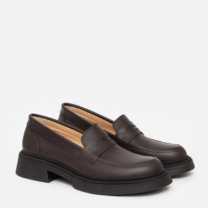 K&ouml;nnte beinhalten: Ein Paar braune Leder-Loafer mit einer klobigen schwarzen Sohle. Die Schuhe haben ein klassisches Penny-Loafer-Design mit runder Spitze und niedrigem Absatz. Das Leder wirkt glatt, vor wei&szlig;em Hintergrund.