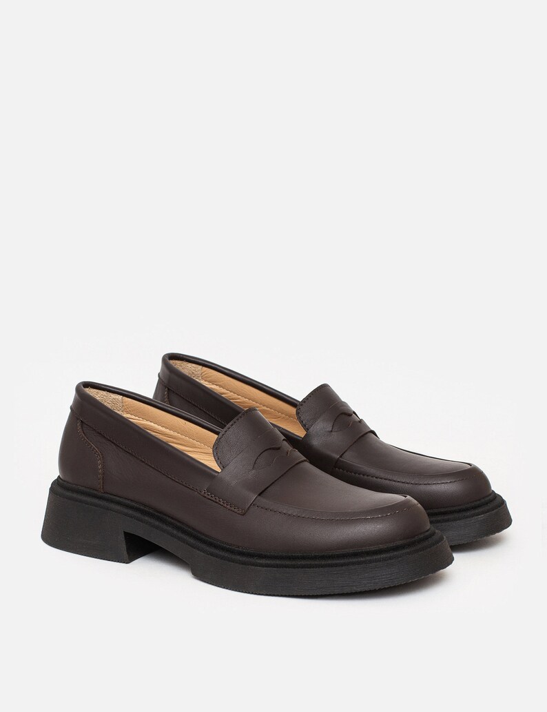 K&ouml;nnte beinhalten: Ein Paar braune Leder-Loafer mit einer klobigen schwarzen Sohle. Die Schuhe haben ein klassisches Penny-Loafer-Design mit runder Spitze und niedrigem Absatz. Das Leder wirkt glatt, vor wei&szlig;em Hintergrund.