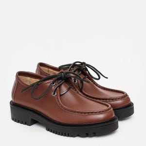 May include: A pair of brown leather shoes featuring a moccasin-style stitched toe, black laces threaded through silver-toned metal eyelets, and a thick, black lugged rubber sole. The shoes have a smooth finish and a rounded toe silhouette.