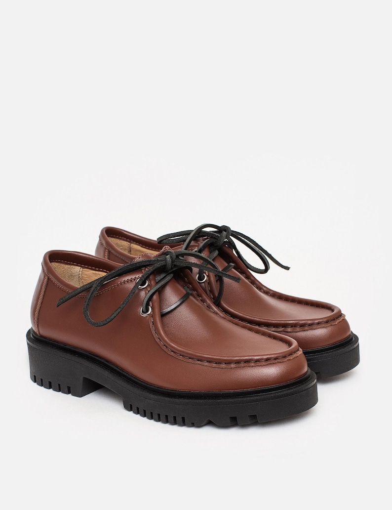 May include: A pair of brown leather shoes featuring a moccasin-style stitched toe, black laces threaded through silver-toned metal eyelets, and a thick, black lugged rubber sole. The shoes have a smooth finish and a rounded toe silhouette.
