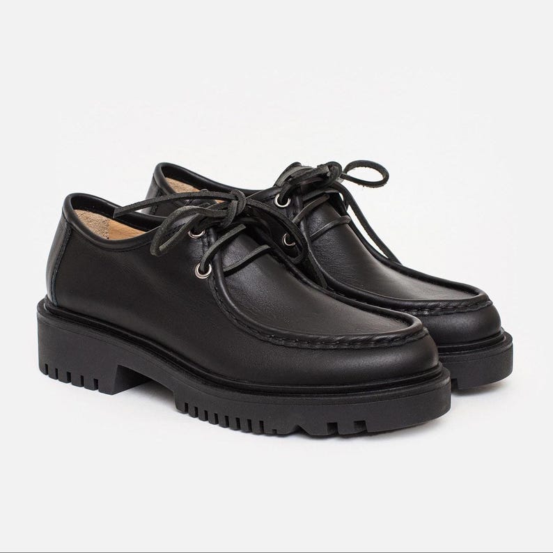May include: A pair of black leather lace-up shoes featuring a moc-toe design with visible stitching along the front edge, silver-toned metal eyelets, thin black laces, and a thick, chunky black rubber lug sole with a deep tread pattern.