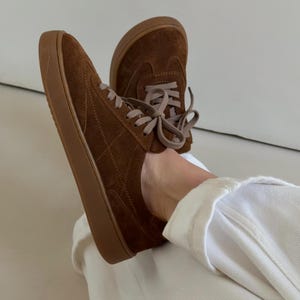 Puede incluir: Zapatillas de ante marr&oacute;n con suelas marrones a juego y cordones marr&oacute;n claro. Los zapatos tienen un patr&oacute;n acolchado en los laterales y se muestran desde un &aacute;ngulo bajo.