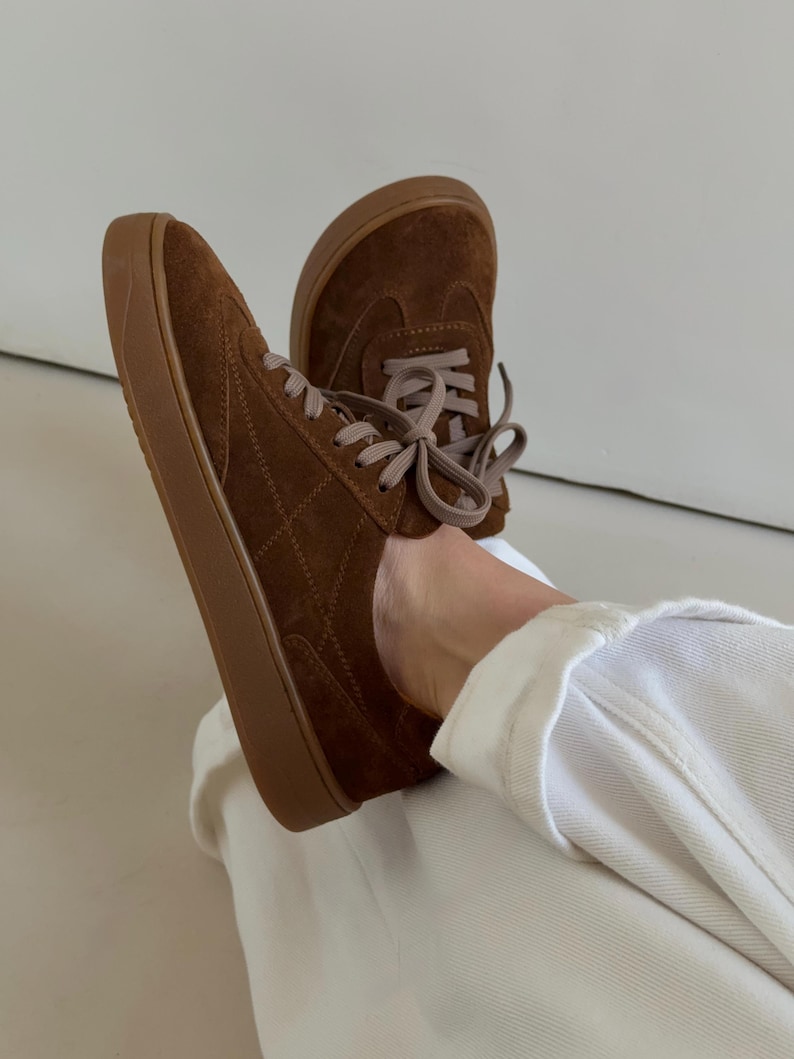 Puede incluir: Zapatillas de ante marr&oacute;n con suelas marrones a juego y cordones marr&oacute;n claro. Los zapatos tienen un patr&oacute;n acolchado en los laterales y se muestran desde un &aacute;ngulo bajo.
