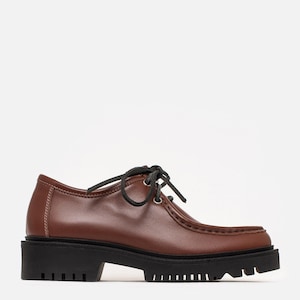 May include: A brown leather oxford shoe featuring a moccasin-style stitched toe, black laces, silver-tone eyelets, and a thick, black lugged rubber sole. The shoe has a low-cut silhouette with visible white stitching along the heel and side panels.