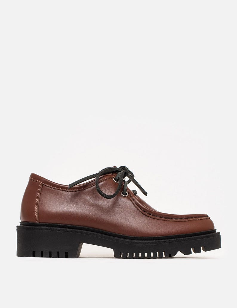 May include: A brown leather oxford shoe featuring a moccasin-style stitched toe, black laces, silver-tone eyelets, and a thick, black lugged rubber sole. The shoe has a low-cut silhouette with visible white stitching along the heel and side panels.