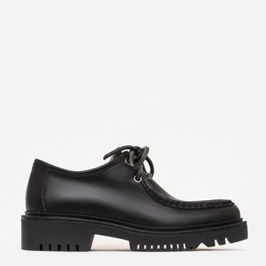 May include: A black leather shoe with a rounded toe, a moccasin-style stitched seam along the front, and a thick, lugged rubber sole. The shoe features a low-cut ankle collar, silver-toned metal eyelets, and black laces tied in a bow at the top.