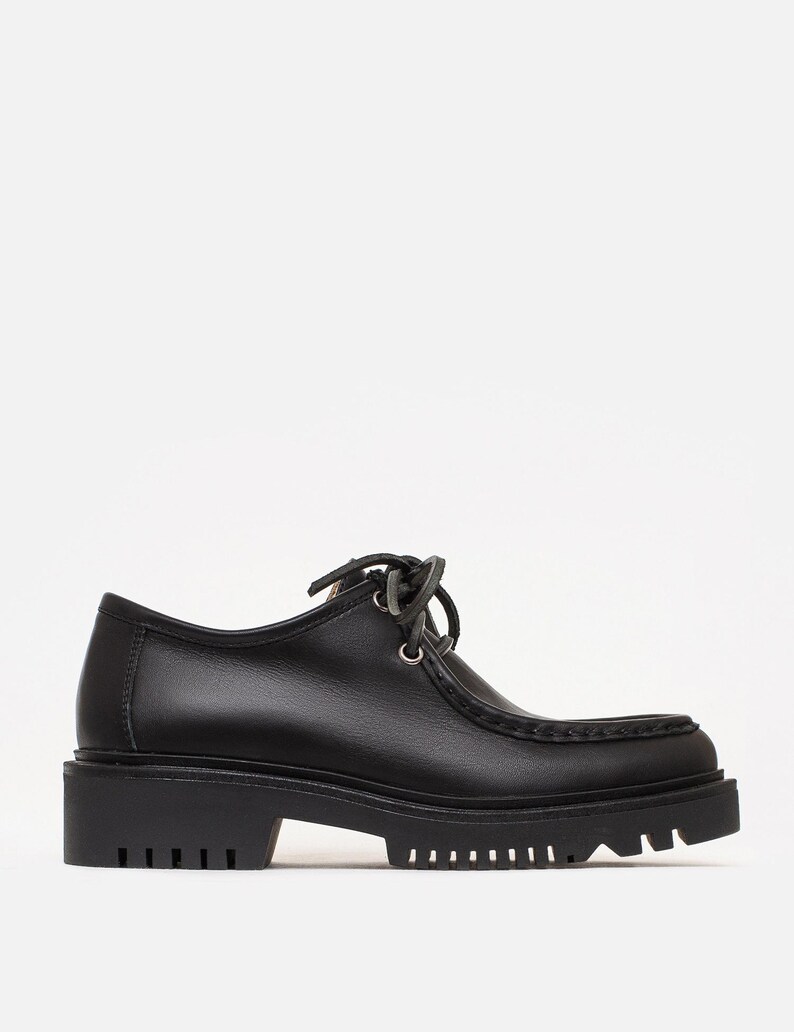 May include: A black leather shoe with a rounded toe, a moccasin-style stitched seam along the front, and a thick, lugged rubber sole. The shoe features a low-cut ankle collar, silver-toned metal eyelets, and black laces tied in a bow at the top.