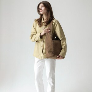 May include: A brown suede hobo bag with a wide shoulder strap and a large exterior pocket, paired with a light tan button-down jacket, white trousers, and dark green slip-on shoes. The bag features a soft, slouchy construction and matte finish.