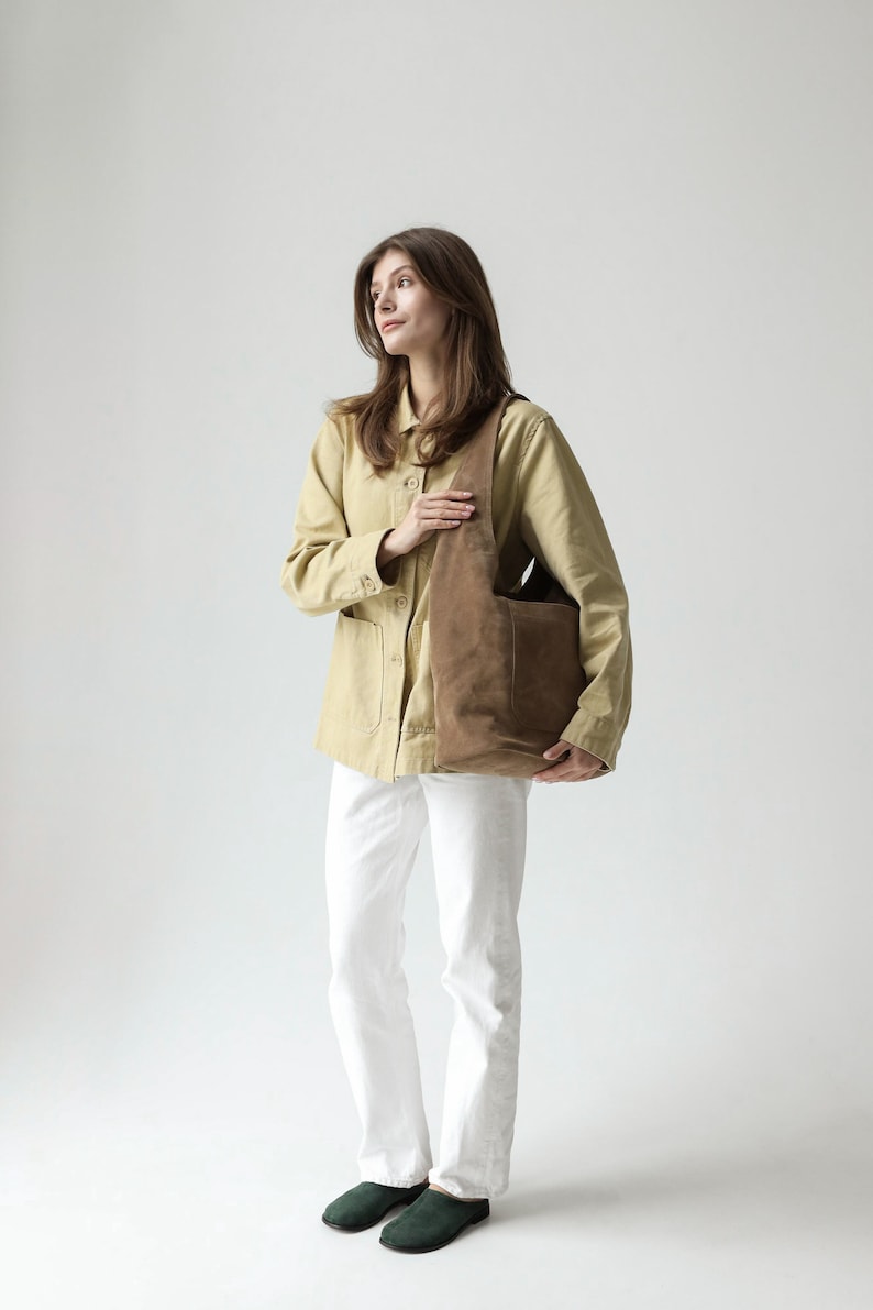May include: A brown suede hobo bag with a wide shoulder strap and a large exterior pocket, paired with a light tan button-down jacket, white trousers, and dark green slip-on shoes. The bag features a soft, slouchy construction and matte finish.