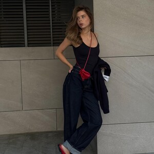 May include: A black tank top, dark wash denim jeans, and red leather clogs. A small red purse hangs from a thin strap. A black jacket is draped over the arm. The outfit is set against a gray wall.