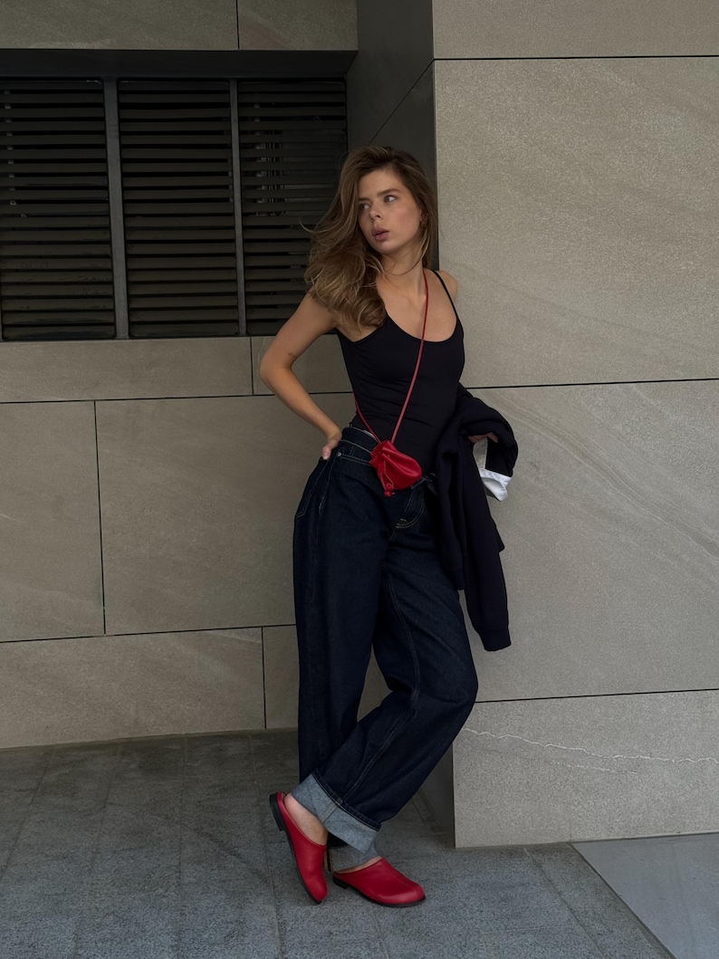 May include: A black tank top, dark wash denim jeans, and red leather clogs. A small red purse hangs from a thin strap. A black jacket is draped over the arm. The outfit is set against a gray wall.