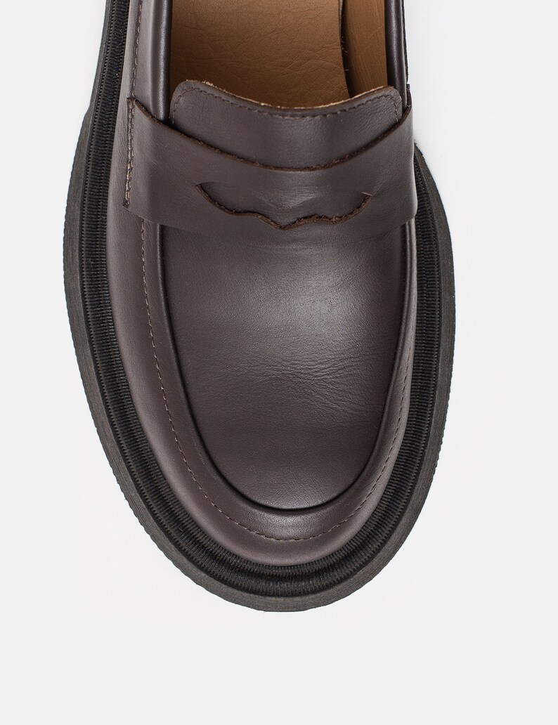 K&ouml;nnte beinhalten: Nahaufnahme eines braunen Lederloafers mit dicker schwarzer Sohle. Der Schuh hat ein klassisches Penny-Loafer-Design mit einem dekorativen Riemen. Die Sohle ist etwa 2,5 cm dick.