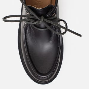 May include: A black leather shoe with a moccasin-style toe, featuring a lace-up closure with thin black laces threaded through silver-toned metal eyelets. The interior lining is tan, with text visible on the insole reading MADE IN UKRAINE.