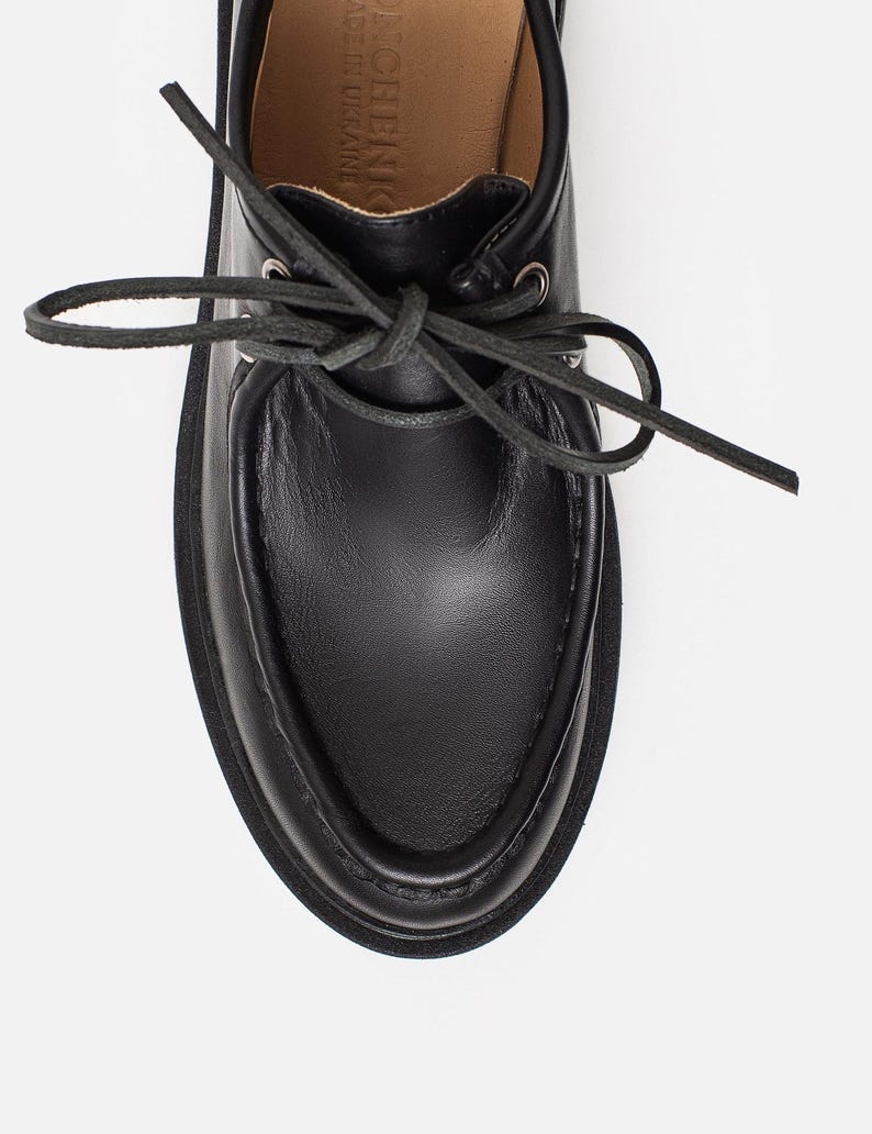May include: A black leather shoe with a moccasin-style toe, featuring a lace-up closure with thin black laces threaded through silver-toned metal eyelets. The interior lining is tan, with text visible on the insole reading MADE IN UKRAINE.
